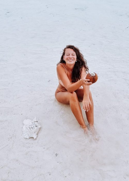 Vrouw zit op een wit strand met een kokosnoot in haar hand