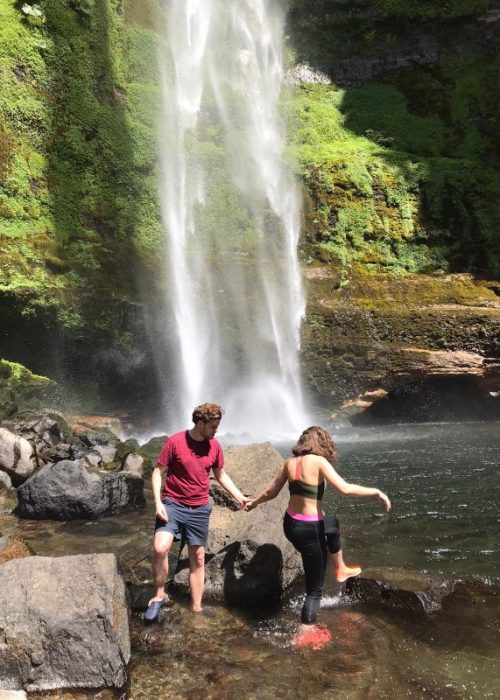 Man en vrouw bij een waterval met waterschoenen aan, vrouw klimt op een rots